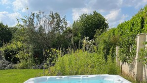 A hot tub with a control panel, surrounded by a wooden deck, set against a backdrop of lush greenery and a clear sky.
