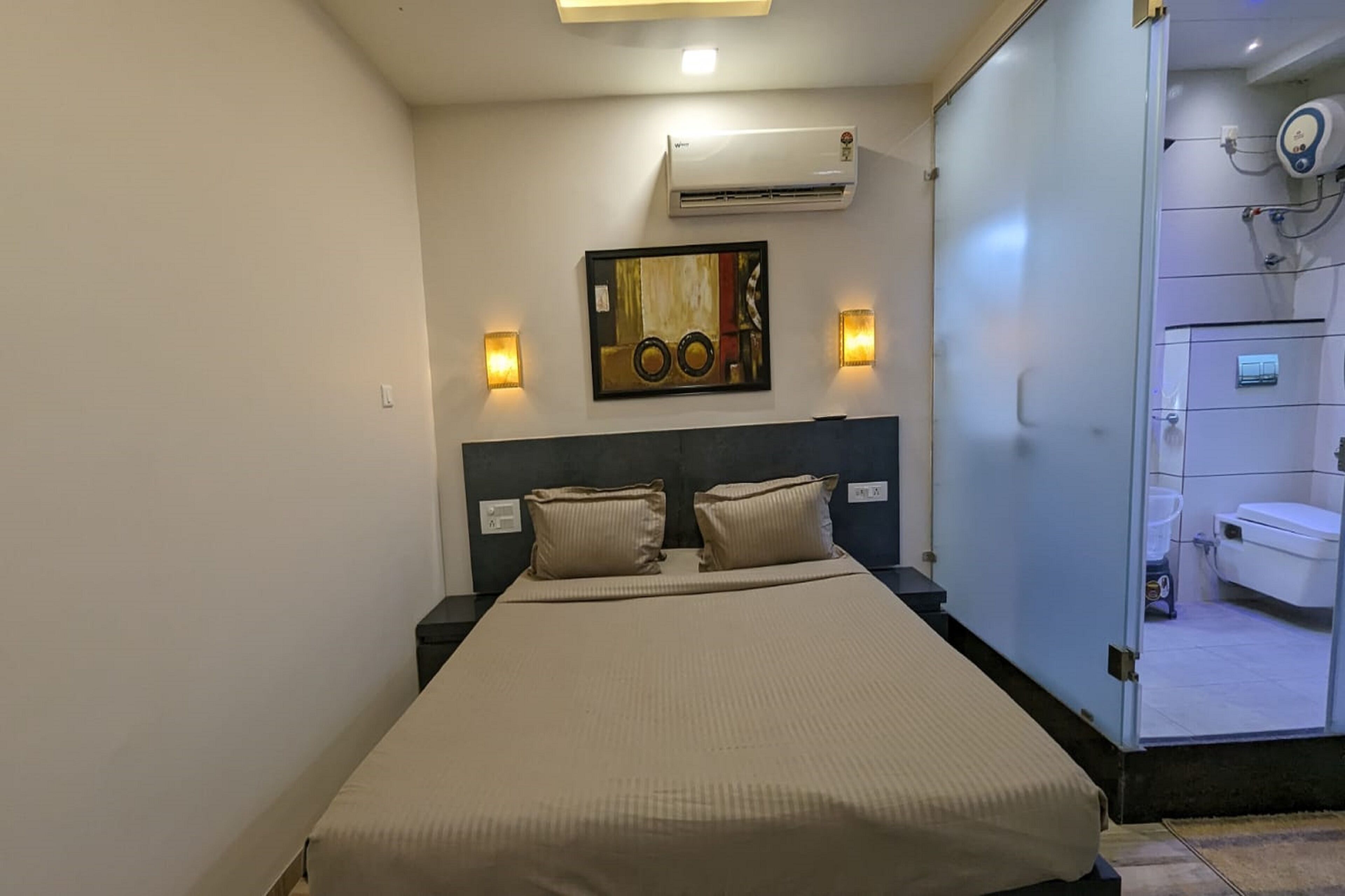 A neatly made bed with a beige bedspread and two pillows, a wall-mounted air conditioner, two wall sconces, and a framed artwork above the bed.