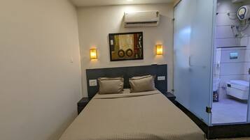 A neatly made bed with a beige bedspread and two pillows, a wall-mounted air conditioner, two wall sconces, and a framed artwork above the bed.