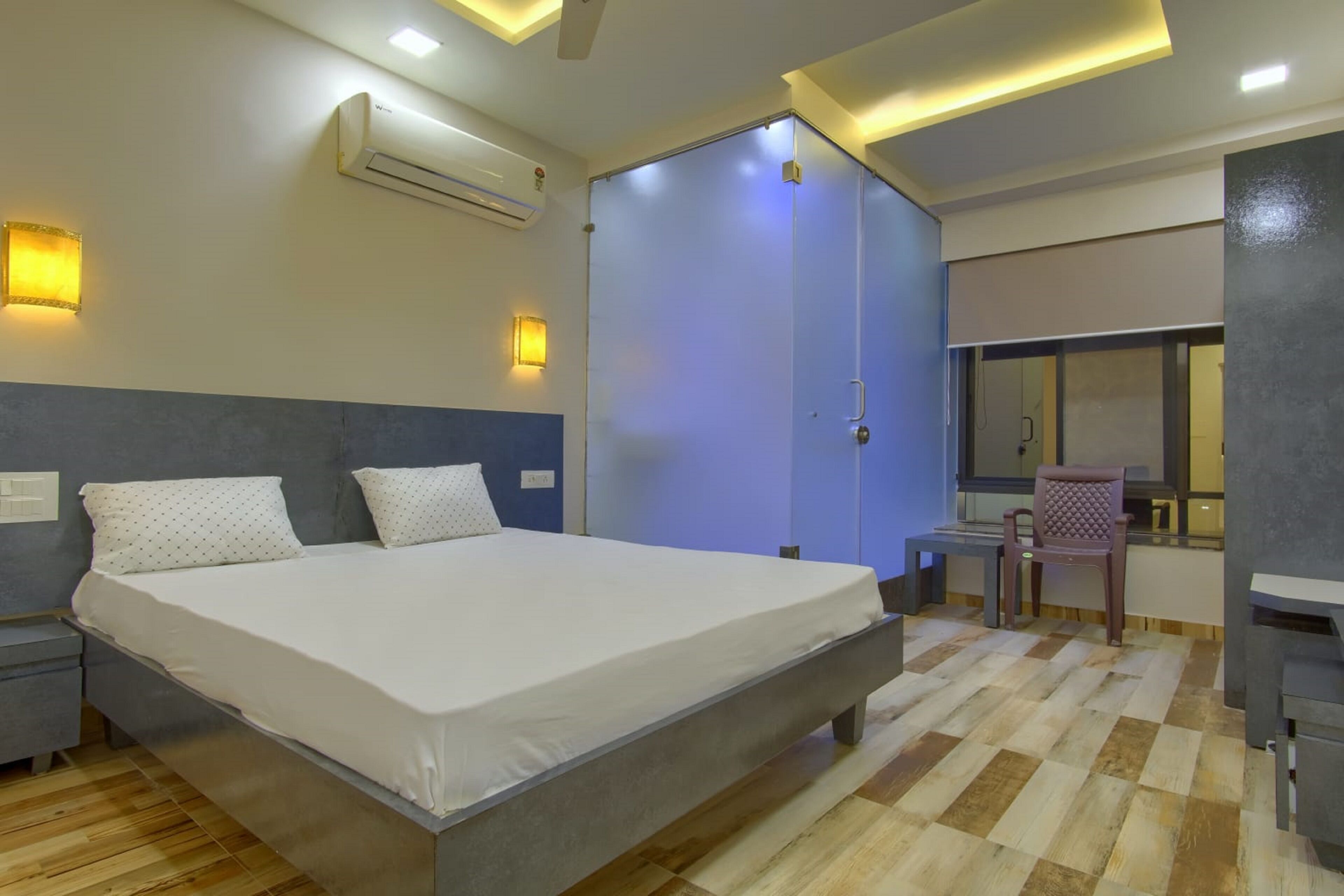 A modern hotel room with a large bed, bedside tables, an air conditioner, a desk with a chair, and a window.
