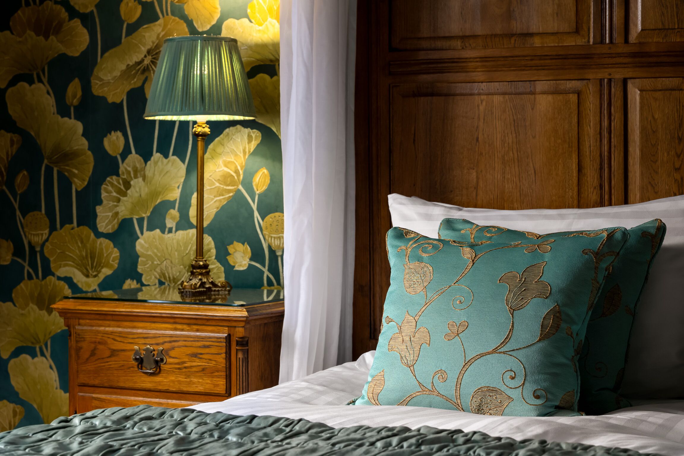 A neatly made bed with a teal pillow and a green lamp on a wooden nightstand.