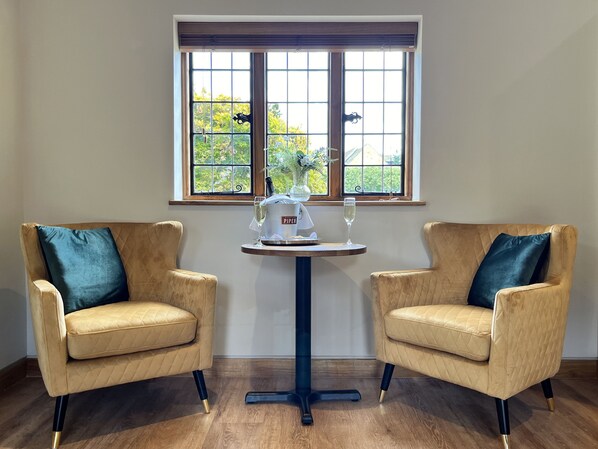 A room with two beige armchairs, a small round table with a white pitcher and two glasses, and a window with a view of greenery.