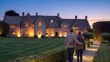 A large stone building with multiple chimneys and windows, surrounded by well-manicured hedges and a gravel path.