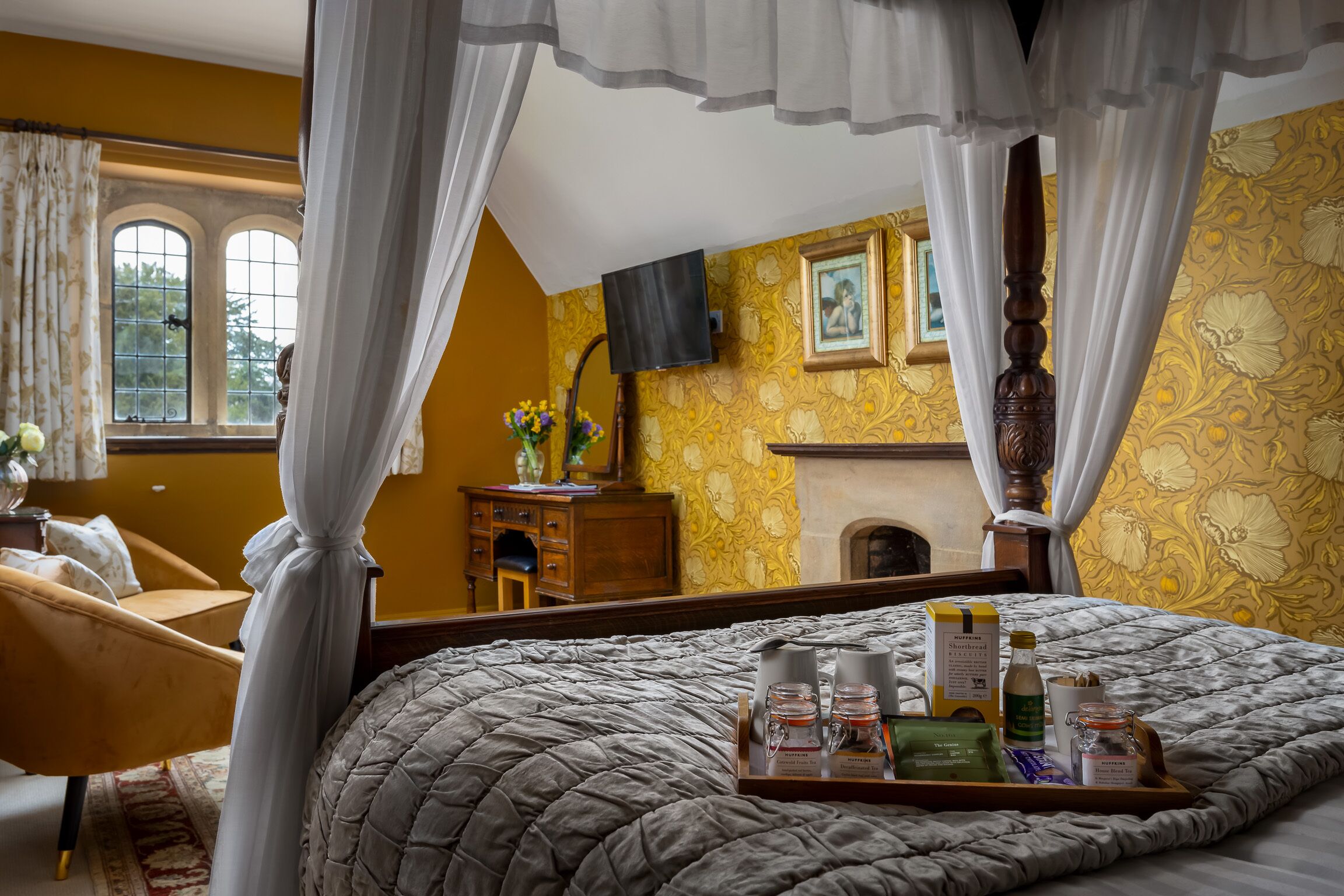 A bedroom with a canopy bed, a bedside table with a tray of breakfast items, a fireplace, and a window with curtains.