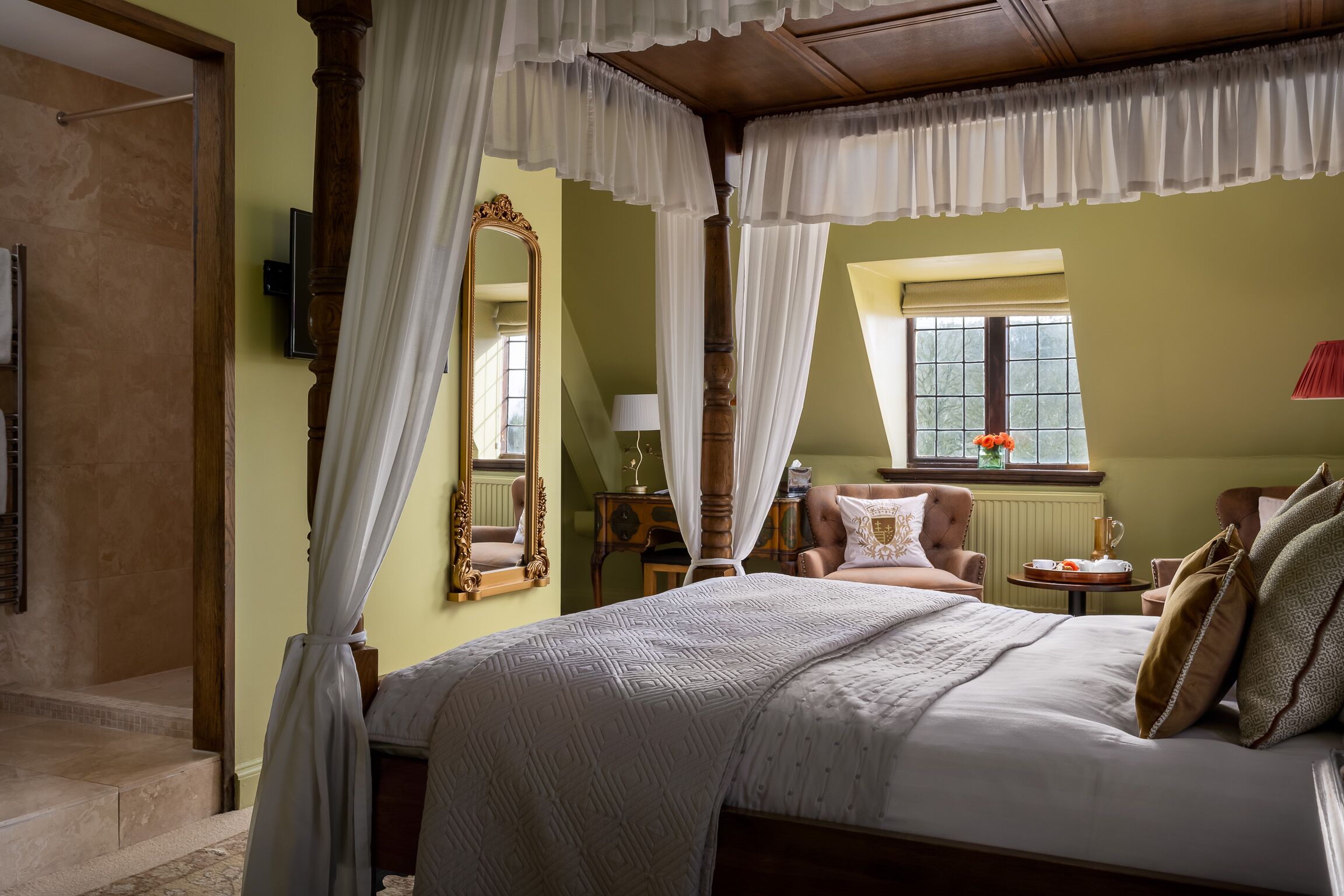 Un dormitorio con cama con dosel, un espejo grande, una silla y una mesita de noche con un jarrón de flores.