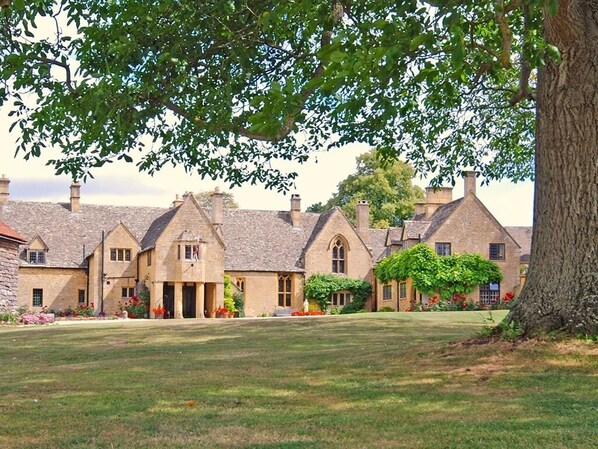 A row of traditional stone buildings with a central entrance, surrounded by a well-maintained lawn and flowering plants. - Abbots Grange Manor House (Broadway)