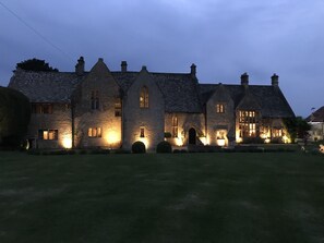 A stone building with multiple chimneys and lit windows, suggesting a traditional architectural style. - Abbots Grange Manor House (Broadway)