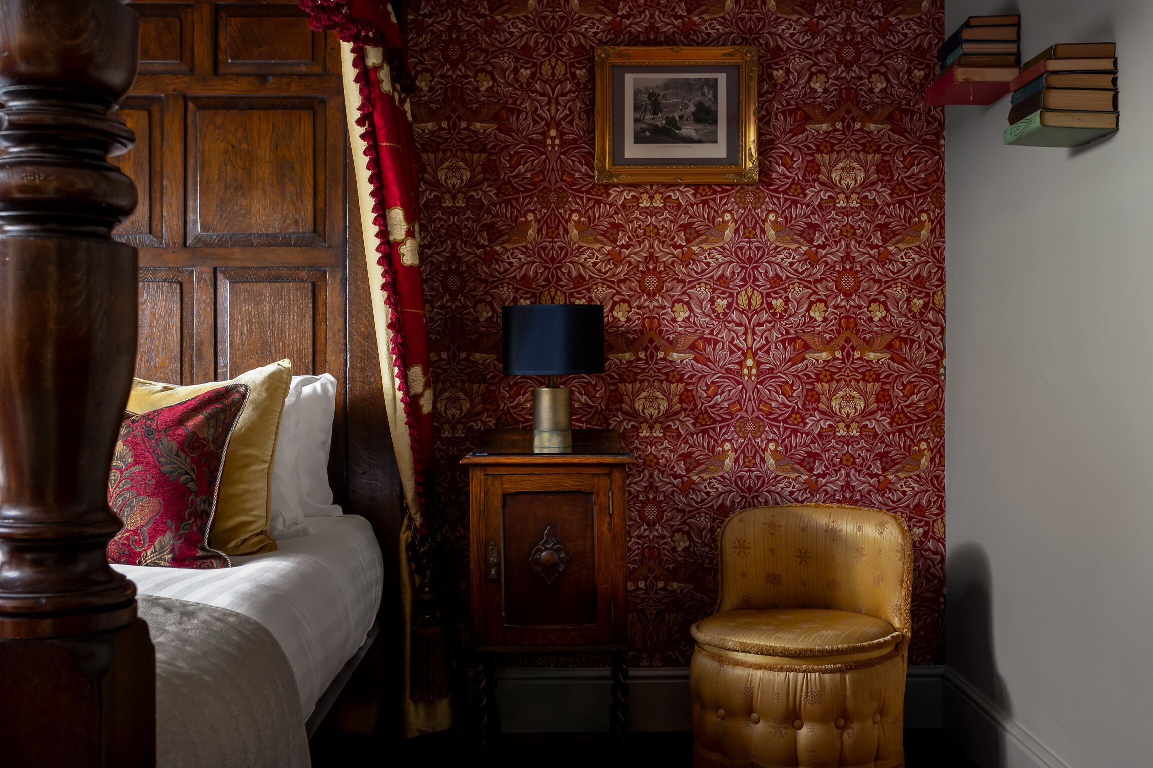 A bedroom with a wooden bed, a red patterned wallpaper, a framed picture, a small wooden cabinet, and a gold armchair.