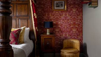 A bedroom with a wooden bed, a red patterned wallpaper, a framed picture, a small wooden cabinet, and a gold armchair.