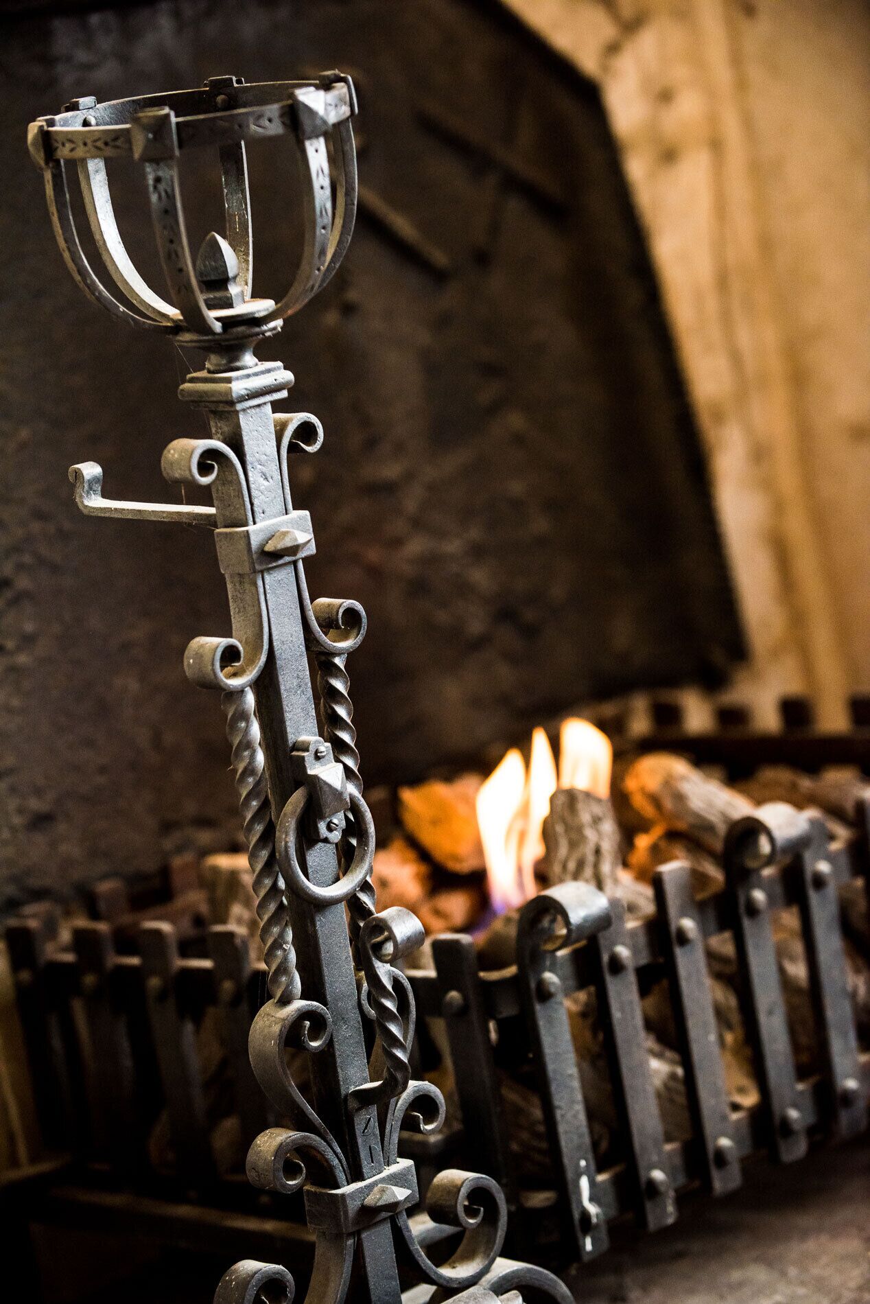 A wrought iron gate with intricate designs and a metal cup at the top, set against a backdrop of a rustic interior with a fire.