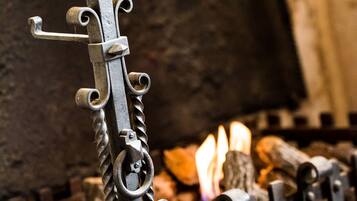A wrought iron gate with intricate designs and a metal cup at the top, set against a backdrop of a rustic interior with a fire.
