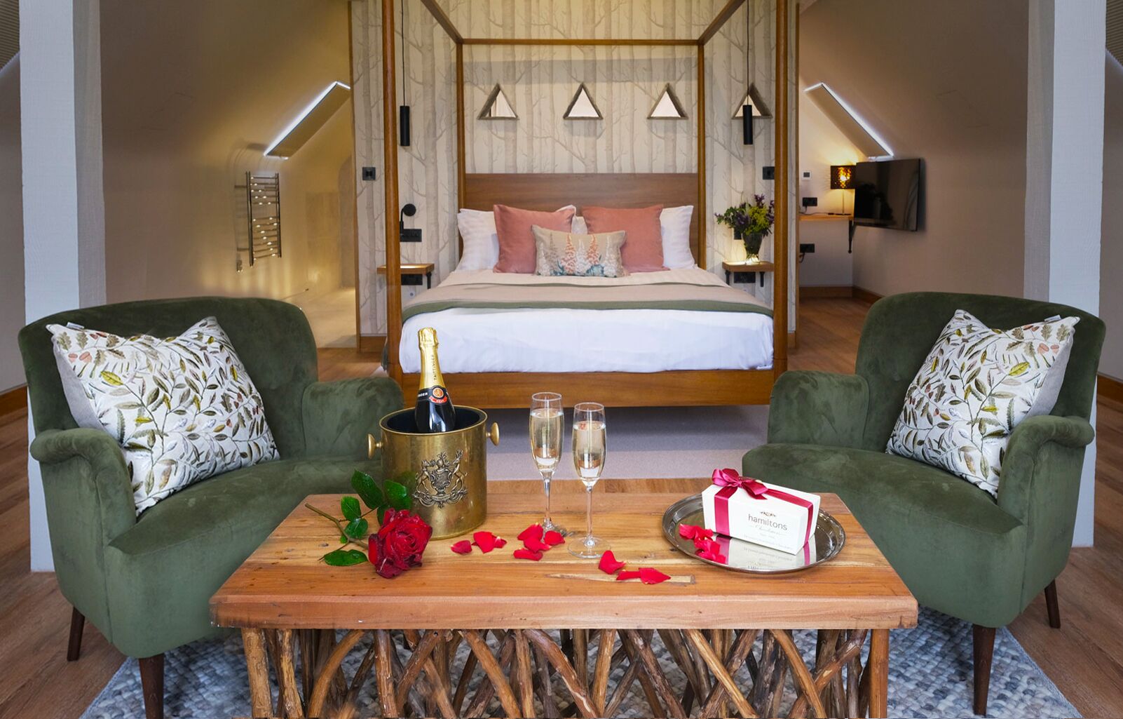 Ett hotellrum med en säng, två gröna fåtöljer, ett träbord med en champagneflaska, glas, en bukett rosor och en presentförpackning.