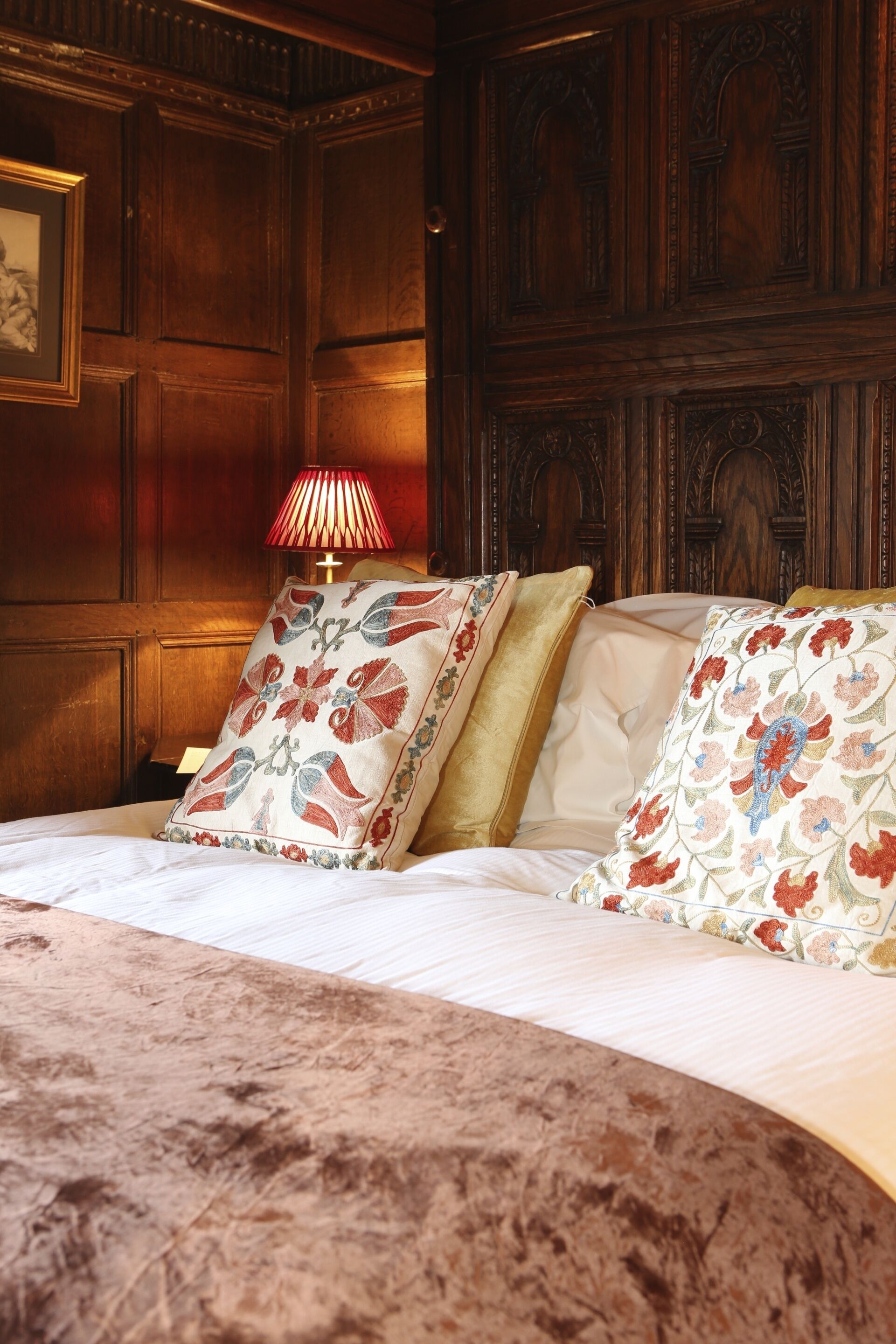 A bedroom with a wooden headboard, a bed with floral-patterned pillows, and a red lamp on a bedside table.