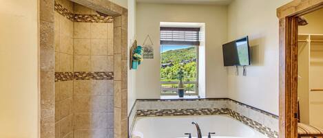 Un baño con una bañera grande, zona de ducha y ventana con vistas a un paisaje verde.