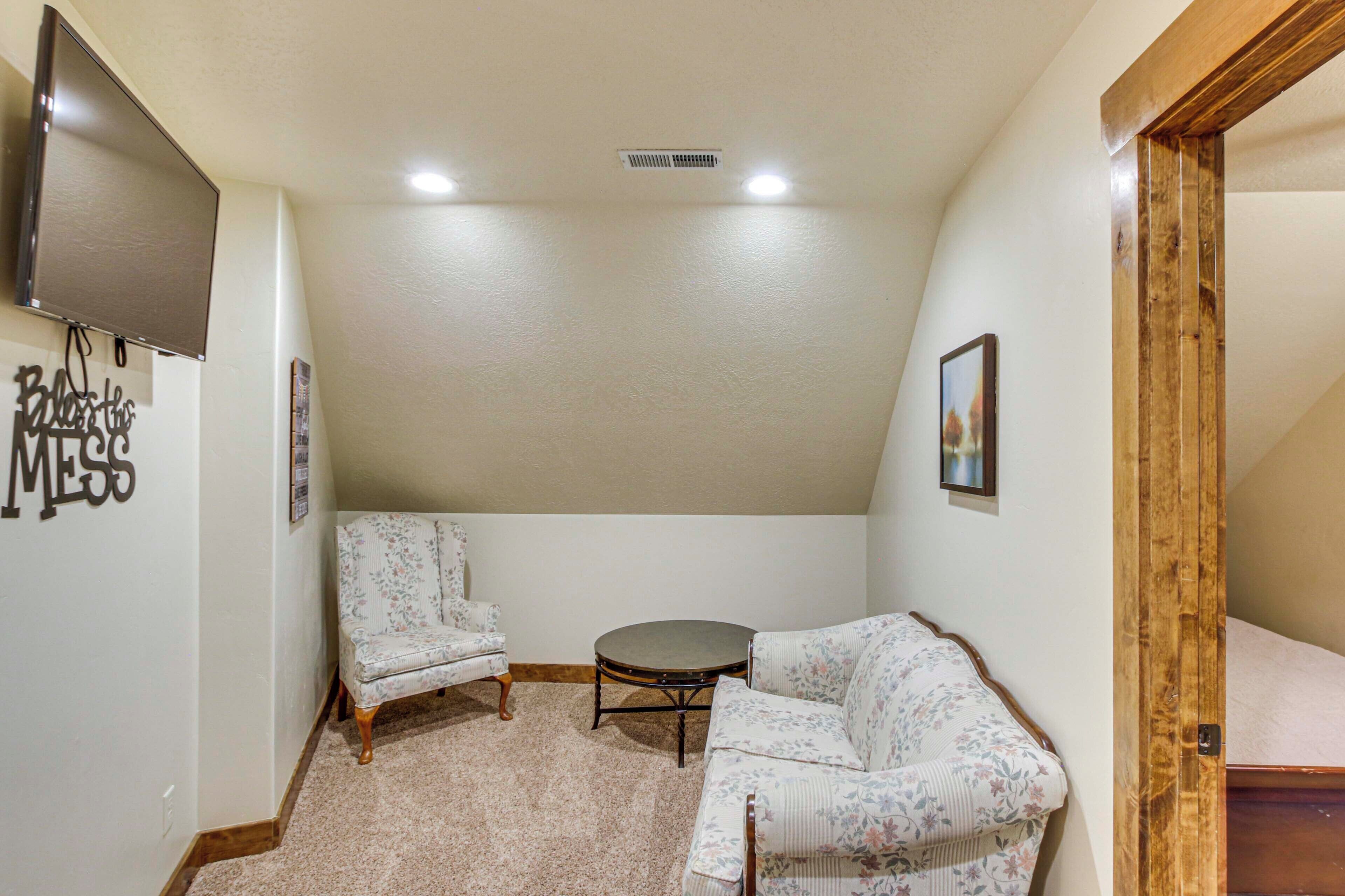 A small, cozy room with a flat-screen TV mounted on the wall, a floral-patterned sofa, a matching armchair, and a small round table.