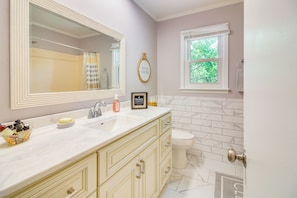 A bathroom with a large mirror, a sink with a soap dish, a framed picture, and a window with a view of trees.