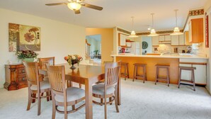Un coin repas avec une table et des chaises en bois, un ventilateur de plafond, et un espace cuisine avec des tabourets et des placards.