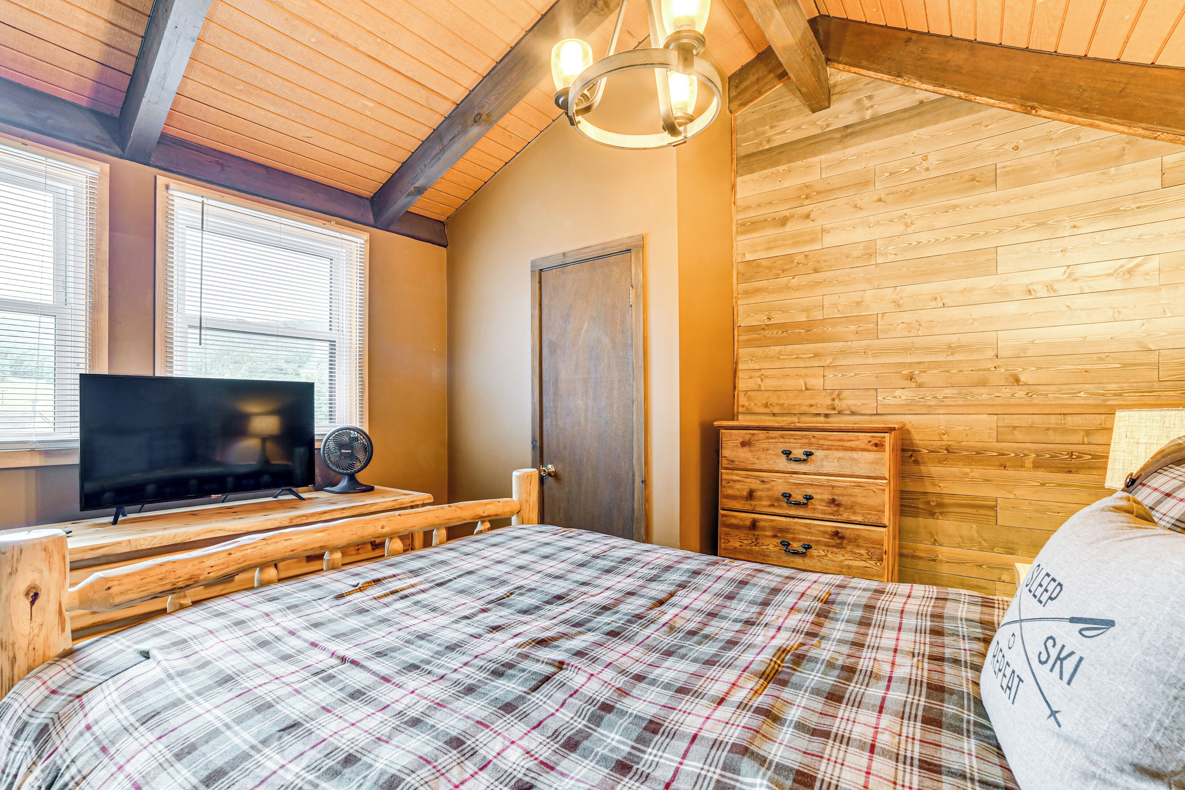 A cozy bedroom with a plaid bedspread, a television, a wooden dresser, and a wooden ceiling.