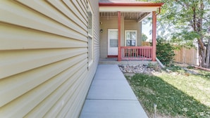A house with a covered porch, a front door, and a small garden area with a wooden fence and trees. - 15 Mi to Downtown Denver: Home w/ Yard + Patio (Aurora)