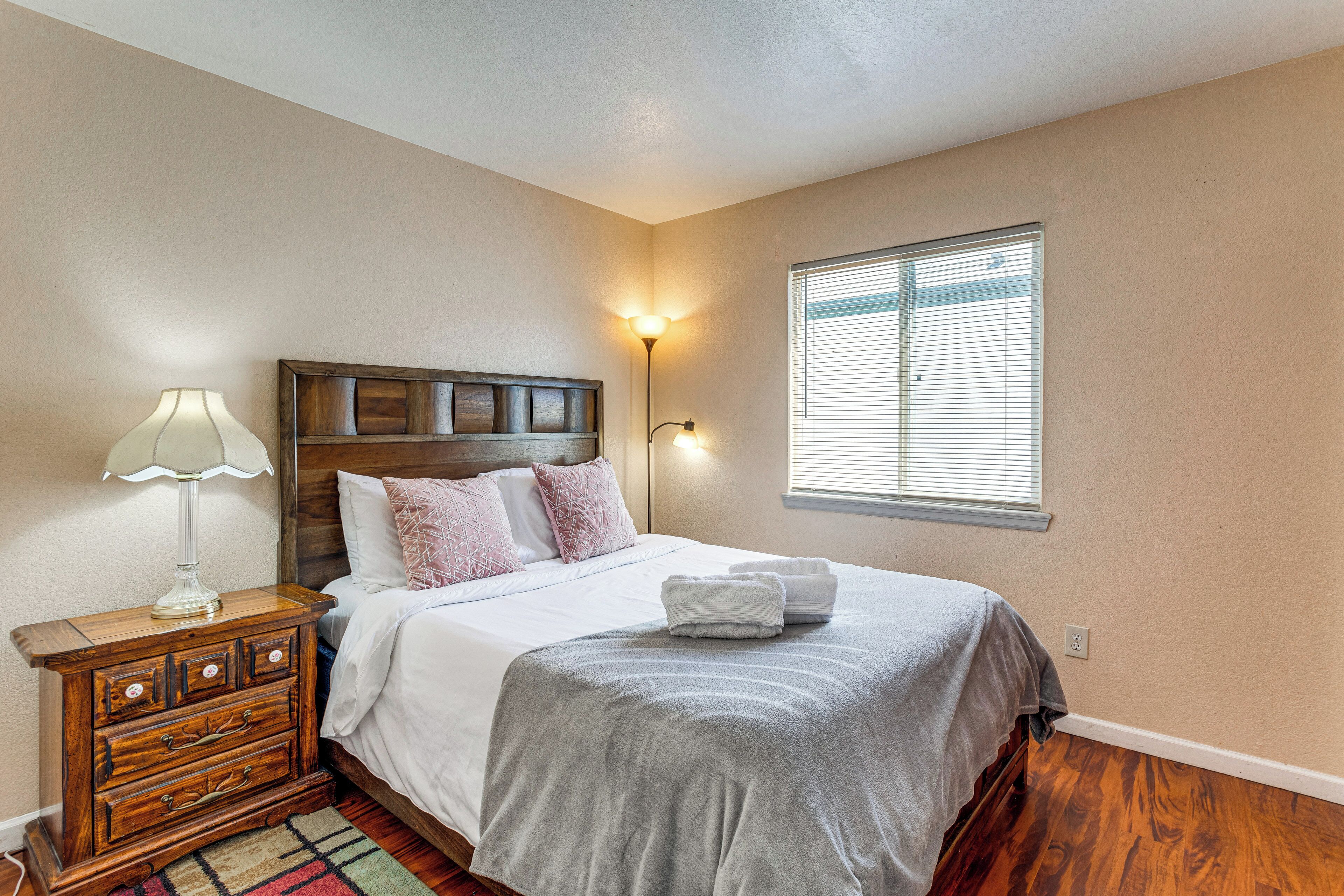 A bedroom with a wooden headboard, a bedside table with a lamp, a bed with a grey blanket, and a window with blinds.