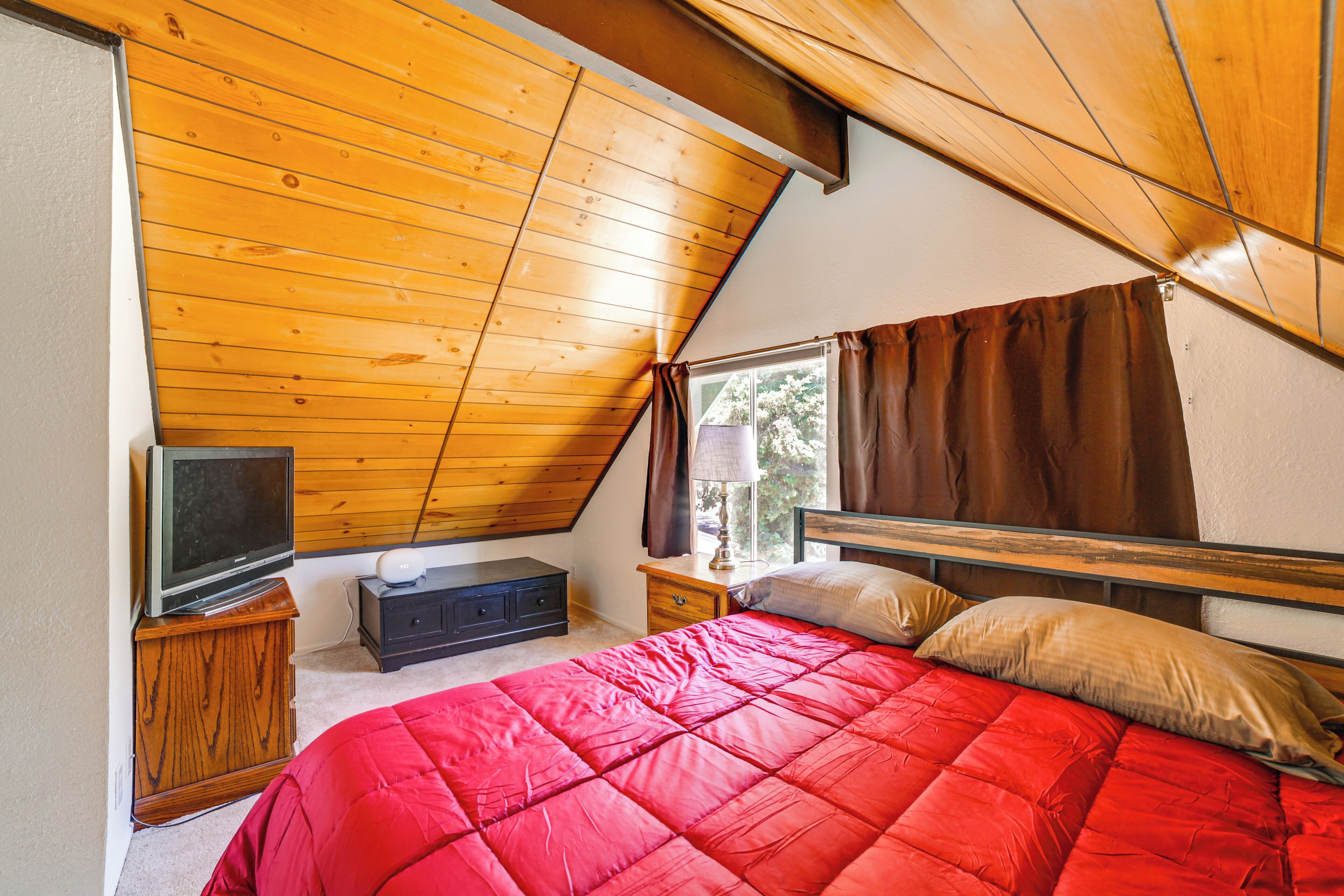 Un dormitorio con techo inclinado, una cama con ropa de cama roja, un televisor sobre un mueble de madera y una ventana con cortinas.