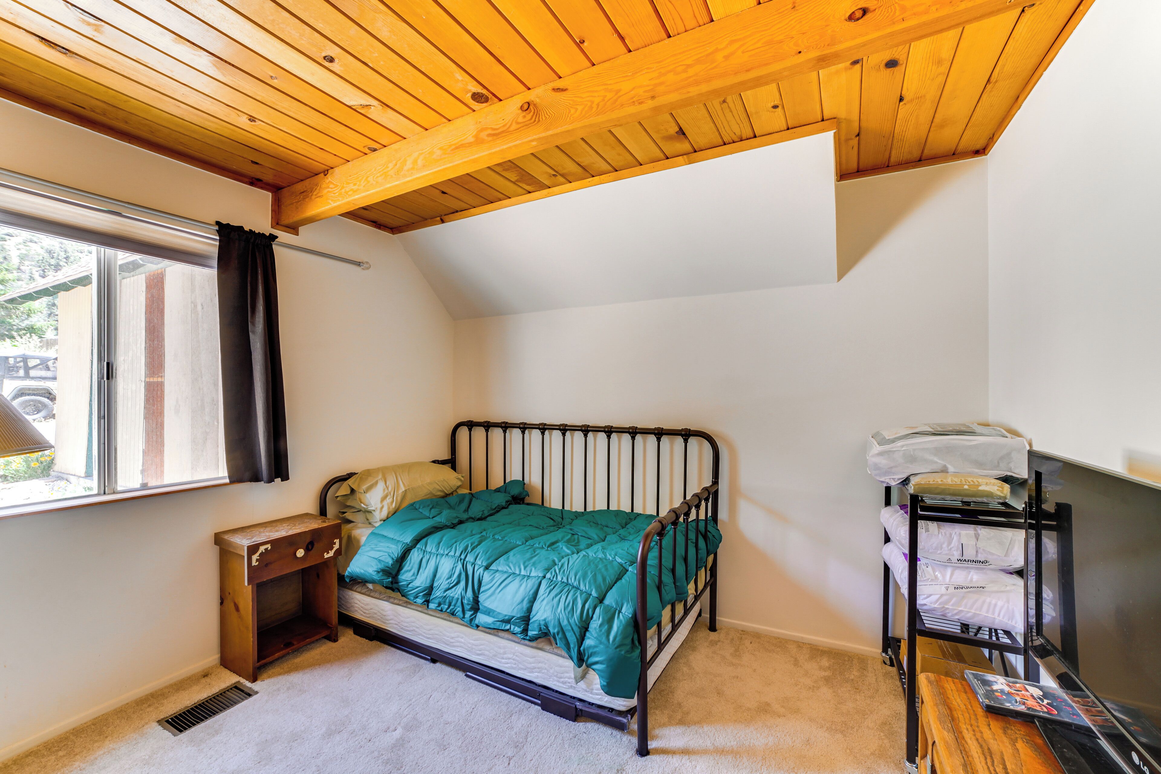 A bedroom with a sloped ceiling, a single bed, a bedside table, a window with curtains, and a small desk with a chair.