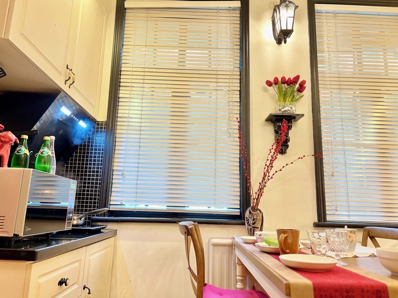 Une cuisine équipée d’un four à micro-ondes, d’une cuisinière et d’une table à manger, cette dernière étant mise avec des assiettes et des verres. On y trouve des fenêtres avec des stores, un luminaire fixé au mur et un vase rempli de fleurs.