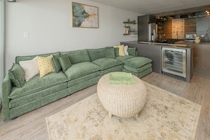 Een groene bank met een geweven poef, een keuken met moderne apparatuur, en een groot abstract schilderij aan de muur.