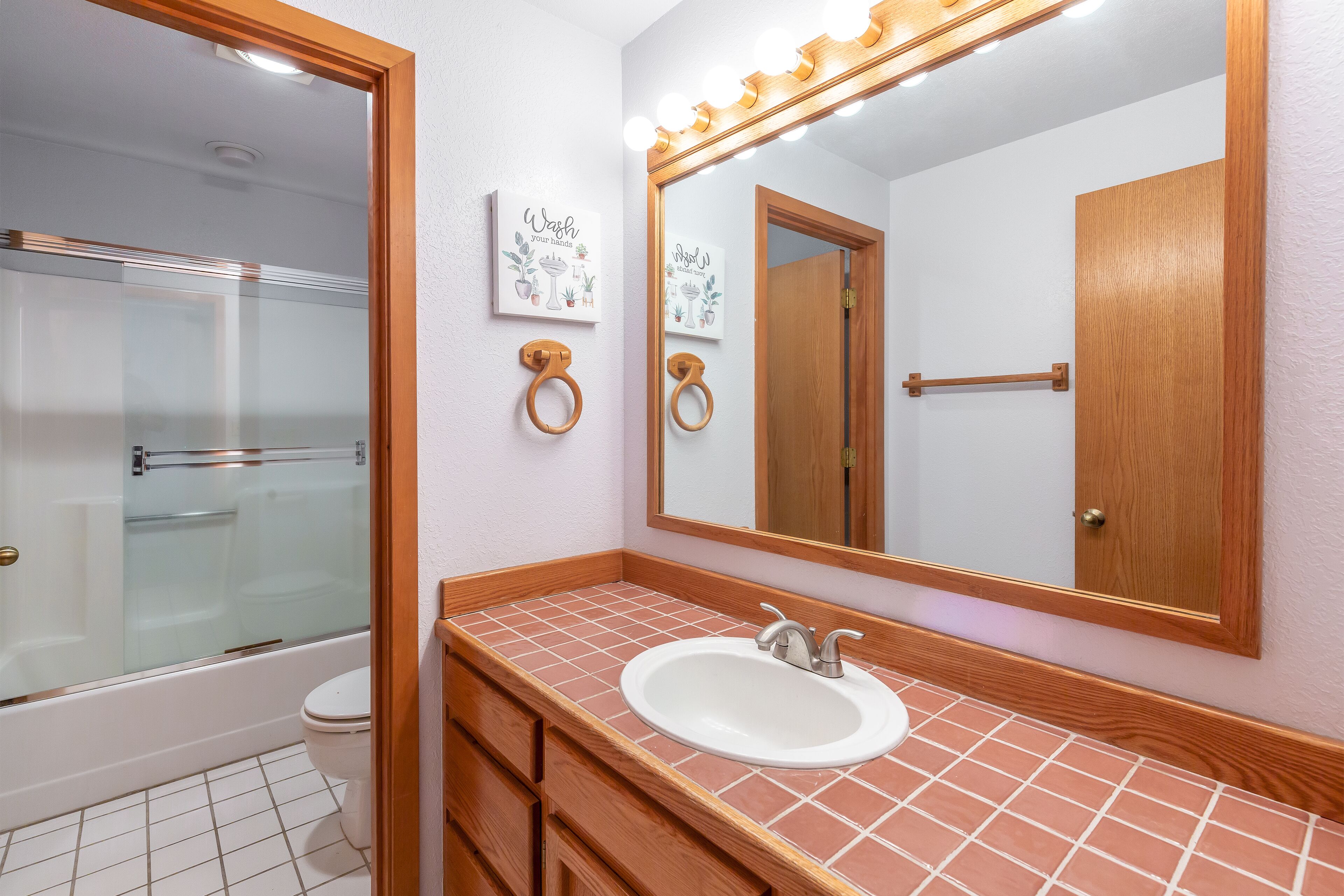 A bathroom with a glass shower enclosure, a white sink with a wooden vanity, and a large mirror with lights above it.