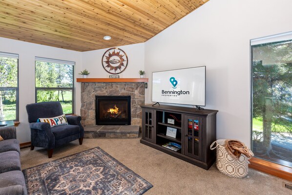 A cozy living room with a stone fireplace, a comfortable armchair, a television, and a large window offering a view of the outdoors. - FAIR27 | Golf Course Views + Private Hot Tub + Bikes | Sleeps 8 (Sunriver)