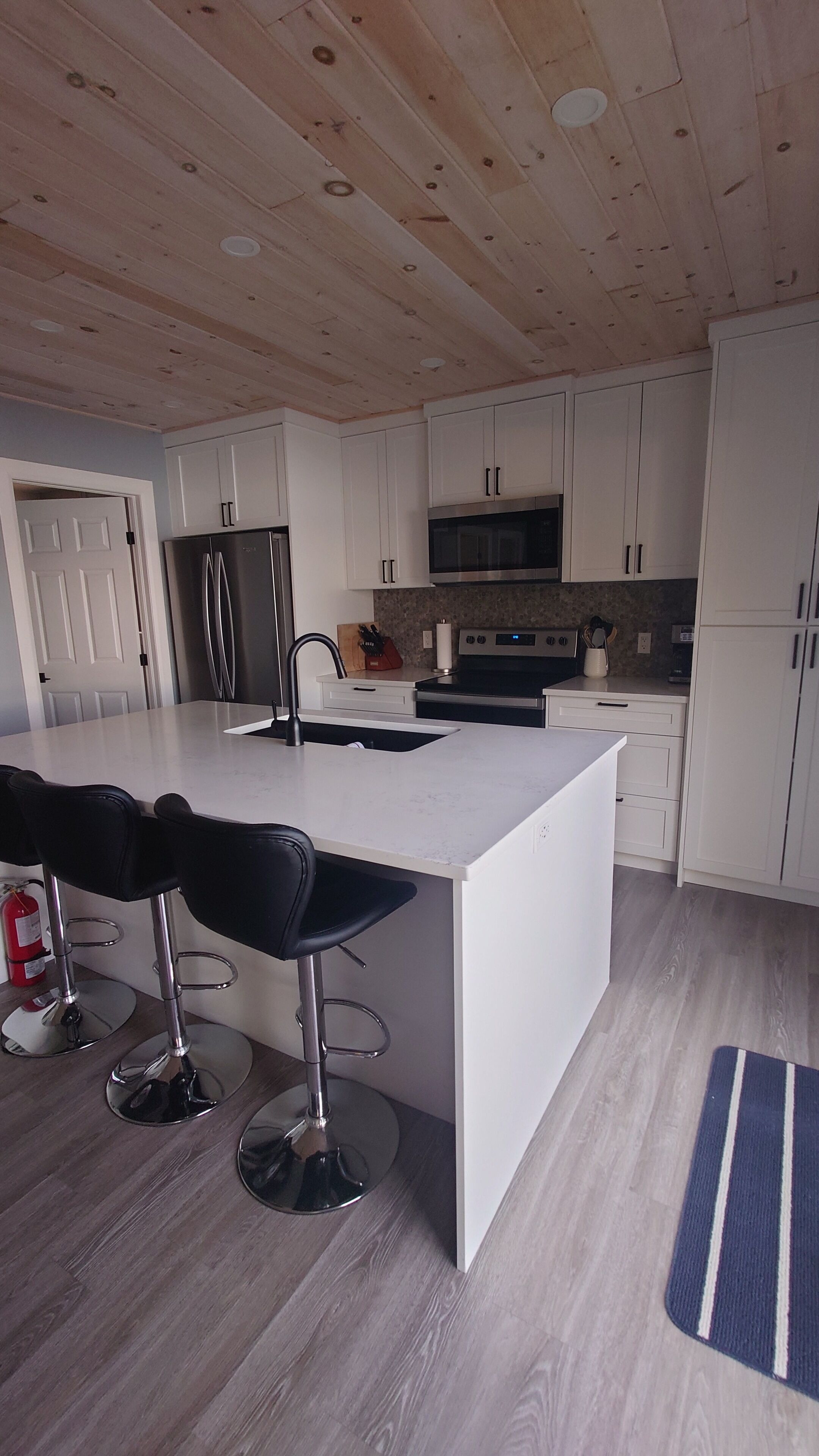 Een moderne keuken met witte kastjes, een centraal eiland met barstoelen, roestvrijstalen apparatuur en een houten plafond.