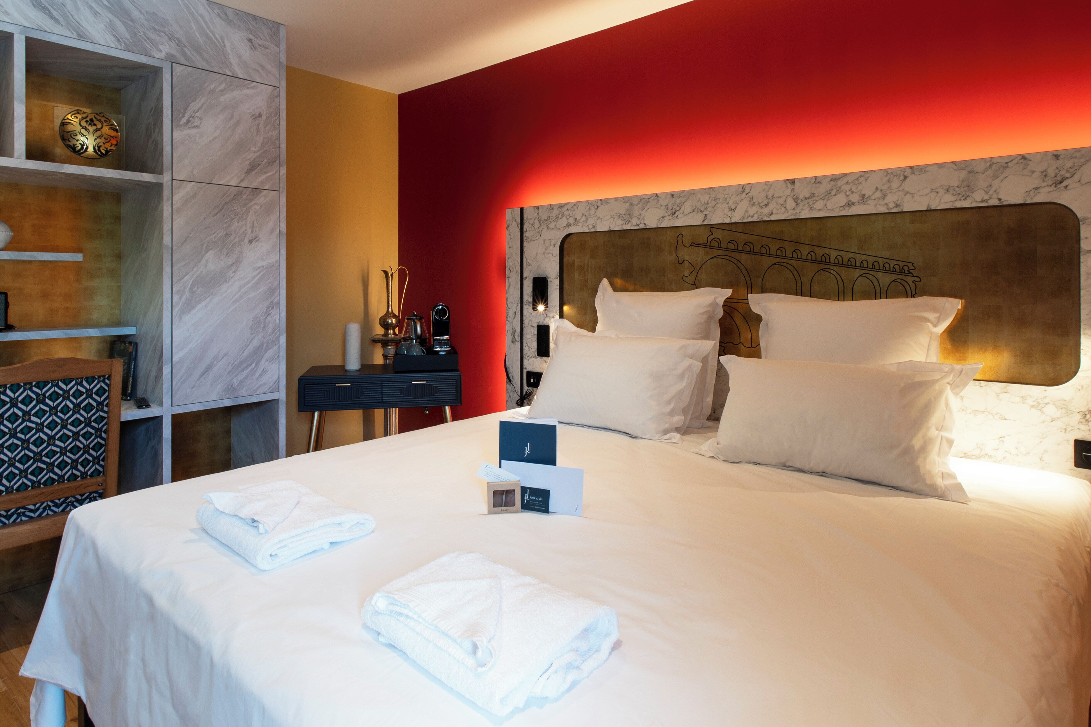 Habitación de hotel con una cama grande, ropa de cama blanca, cabecera de mármol y una pared de acento roja.