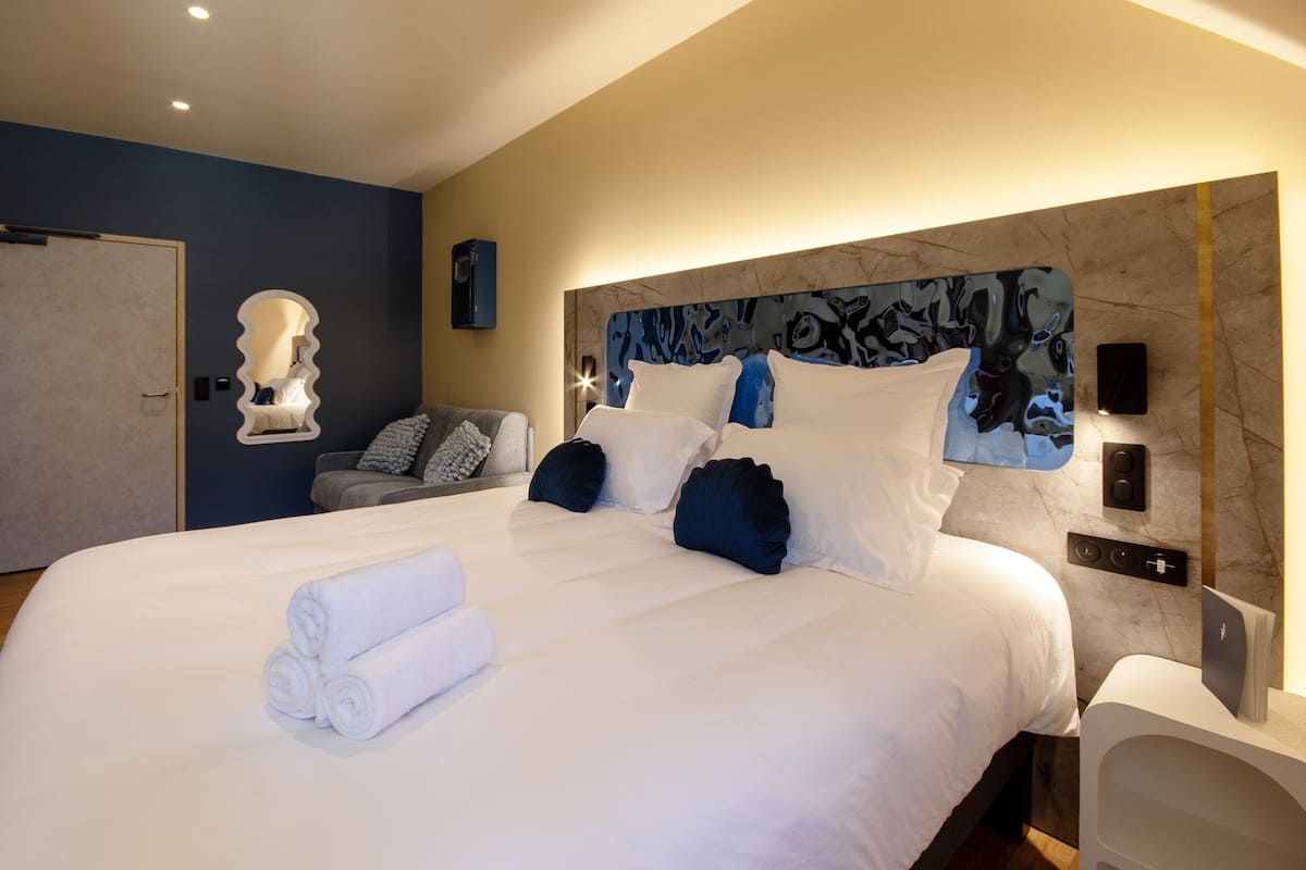 A modern hotel room with a large bed, a headboard with a blue abstract design, a mirror, and a seating area with a sofa and armchairs.
