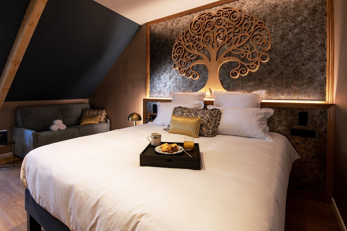 A modern bedroom with a large bed, a decorative tree wall art, a sofa with cushions, and a tray with breakfast items.