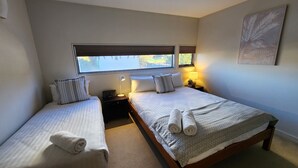 A hotel room with two single beds, a nightstand with a lamp, a window with blinds, and a wall-mounted artwork.