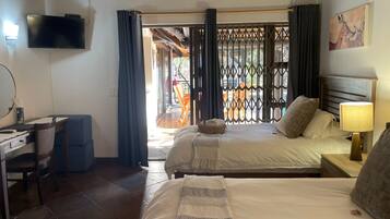 A hotel room with a bed, bedside table, lamp, desk with chair, TV, and a view through a window with curtains.