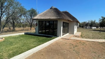 A small building with a thatched roof and large windows, surrounded by a grassy area and trees.