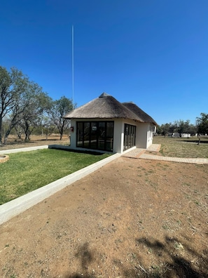 A small building with a thatched roof and large windows, surrounded by a grassy area and trees. - Halali Wilderness Game Ranch (Hammanskraal)