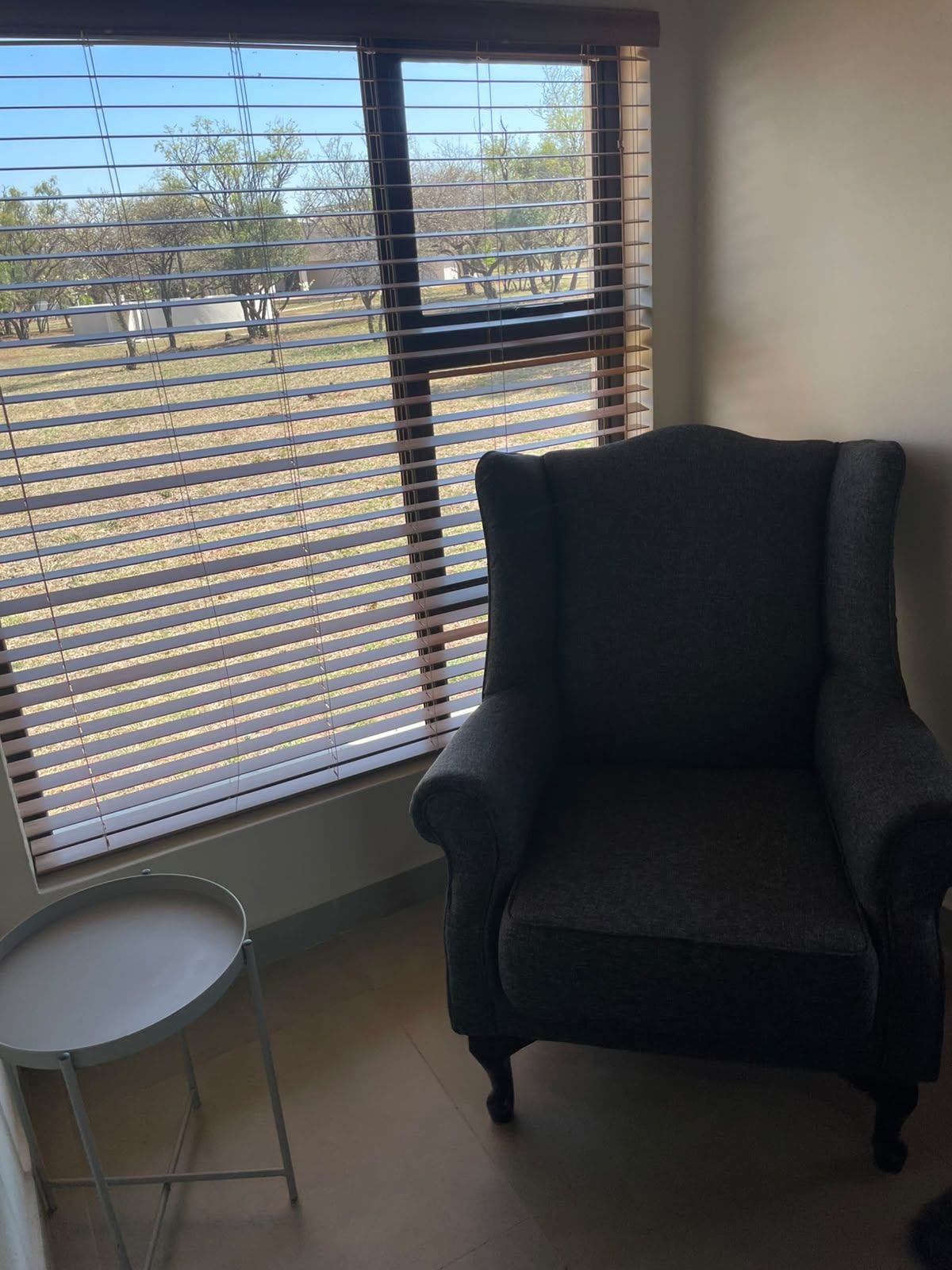 Een kamer met een donker fauteuil, een klein rond tafeltje en een raam met jaloezieën.