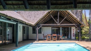 A pool area with a thatched roof structure, a table with chairs, and a view of the surrounding landscape.