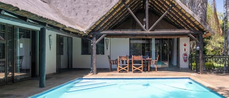 A pool area with a thatched roof structure, a table with chairs, and a view of the surrounding landscape.