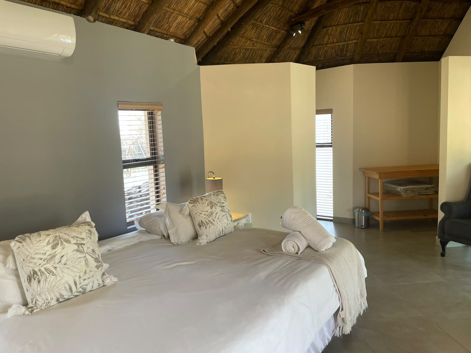 A bedroom with a bed, pillows, a lamp, a wooden table, and a trash bin.