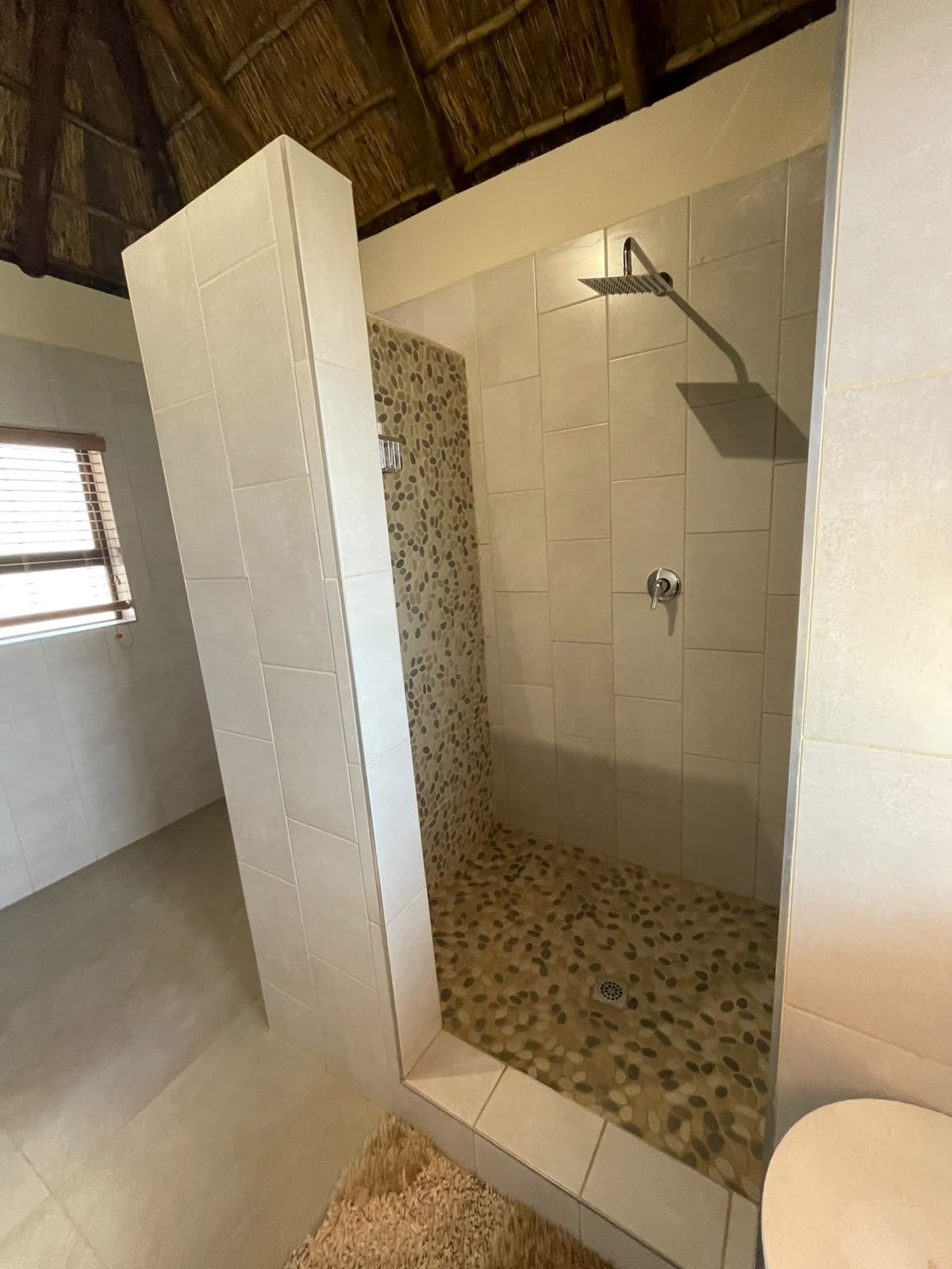 A shower with a unique pebble-patterned wall, a white tiled floor, and a rustic wooden ceiling.