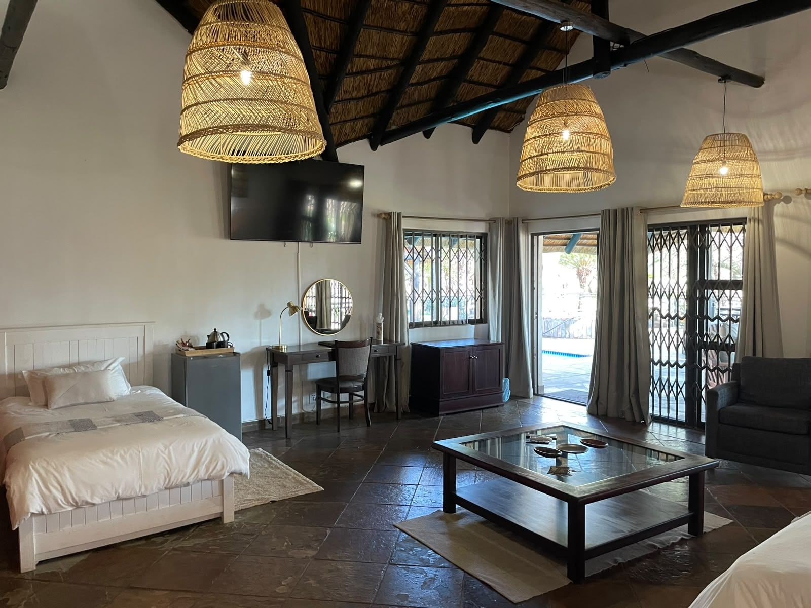 A room with a bed, desk, and seating area, featuring a TV, wicker lamps, and a glass coffee table.