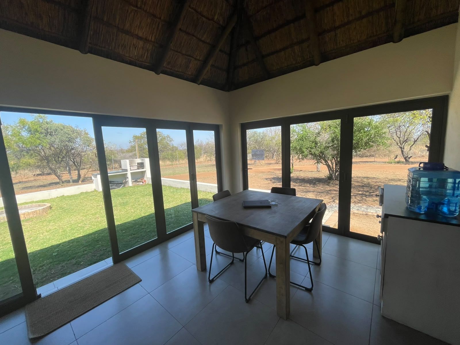 Una habitación con una mesa de comedor y sillas, ventanas amplias con vista a un área verde y árboles, y un dispensador de agua sobre un mueble.
