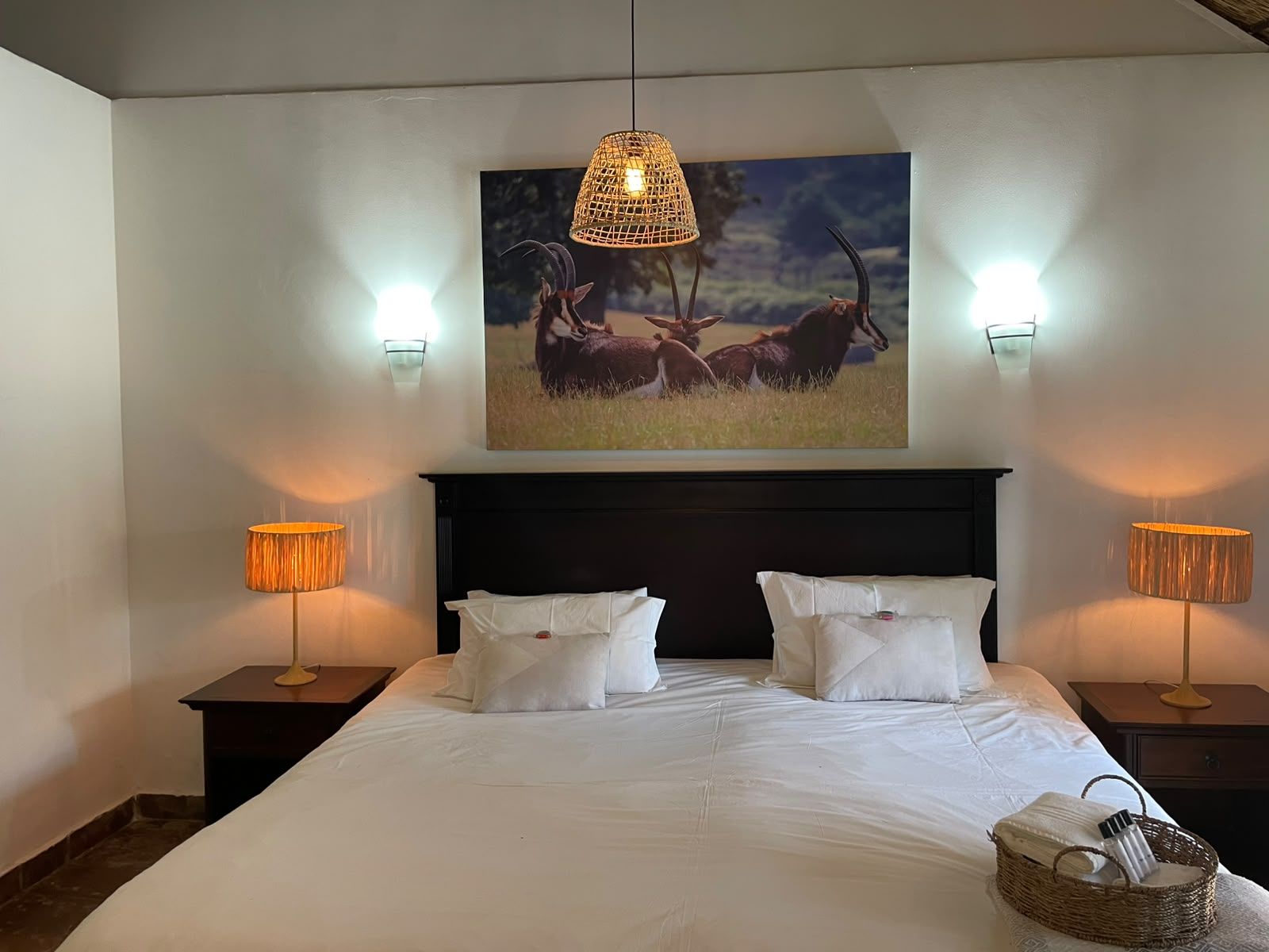 A bedroom with a large bed, two bedside lamps, a wall-mounted picture of antelopes, and a woven basket on the bed.
