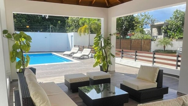 A covered outdoor seating area with wicker furniture and a pool in the background. - Ático de lujos  (Santiago de los Caballeros)