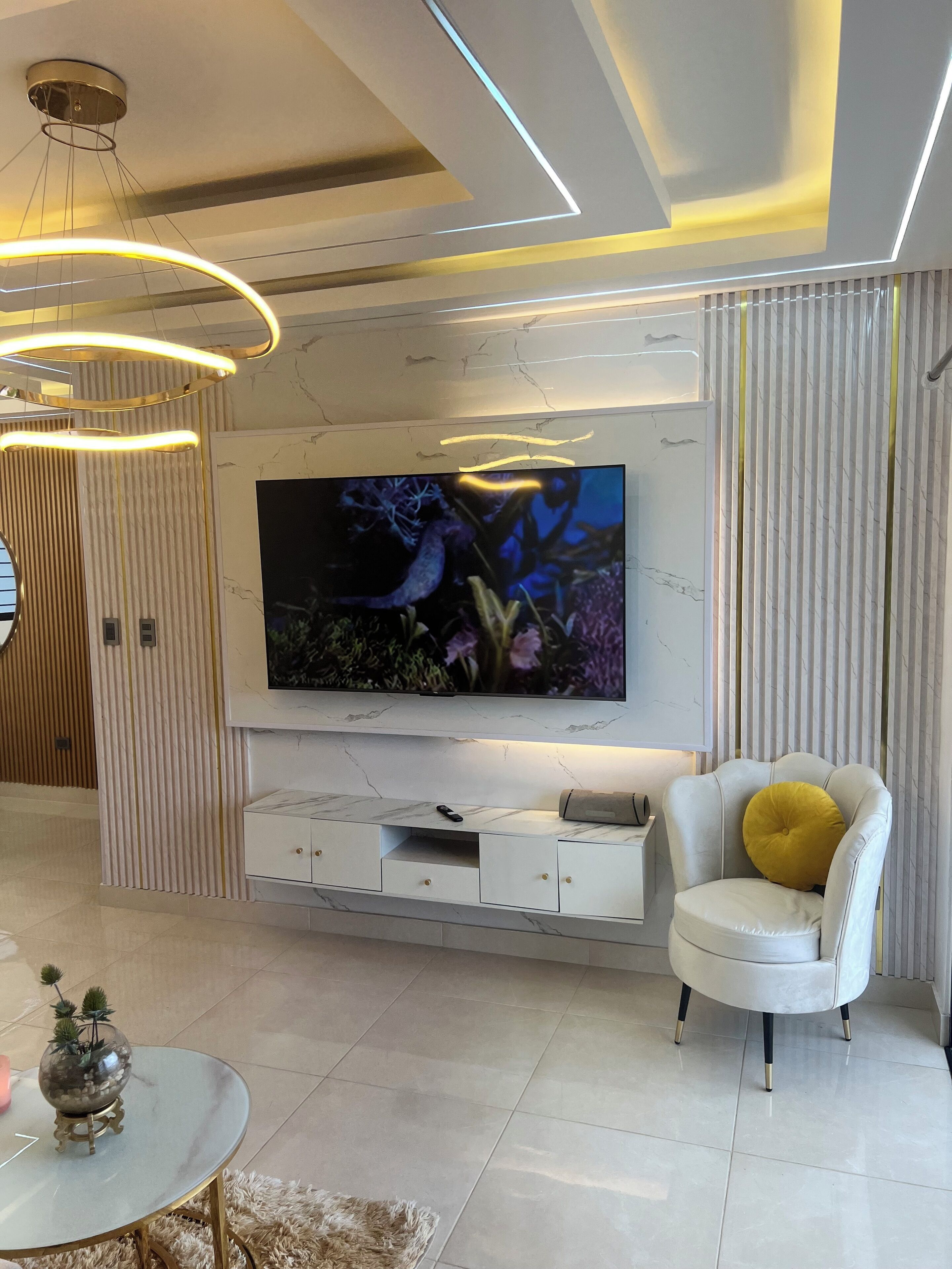 Un salon moderne avec un grand téléviseur à écran plat fixé sur un mur en marbre, un fauteuil en cuir blanc agrémenté d’un coussin jaune et une table basse ronde en verre sur laquelle est posée une plante décorative.