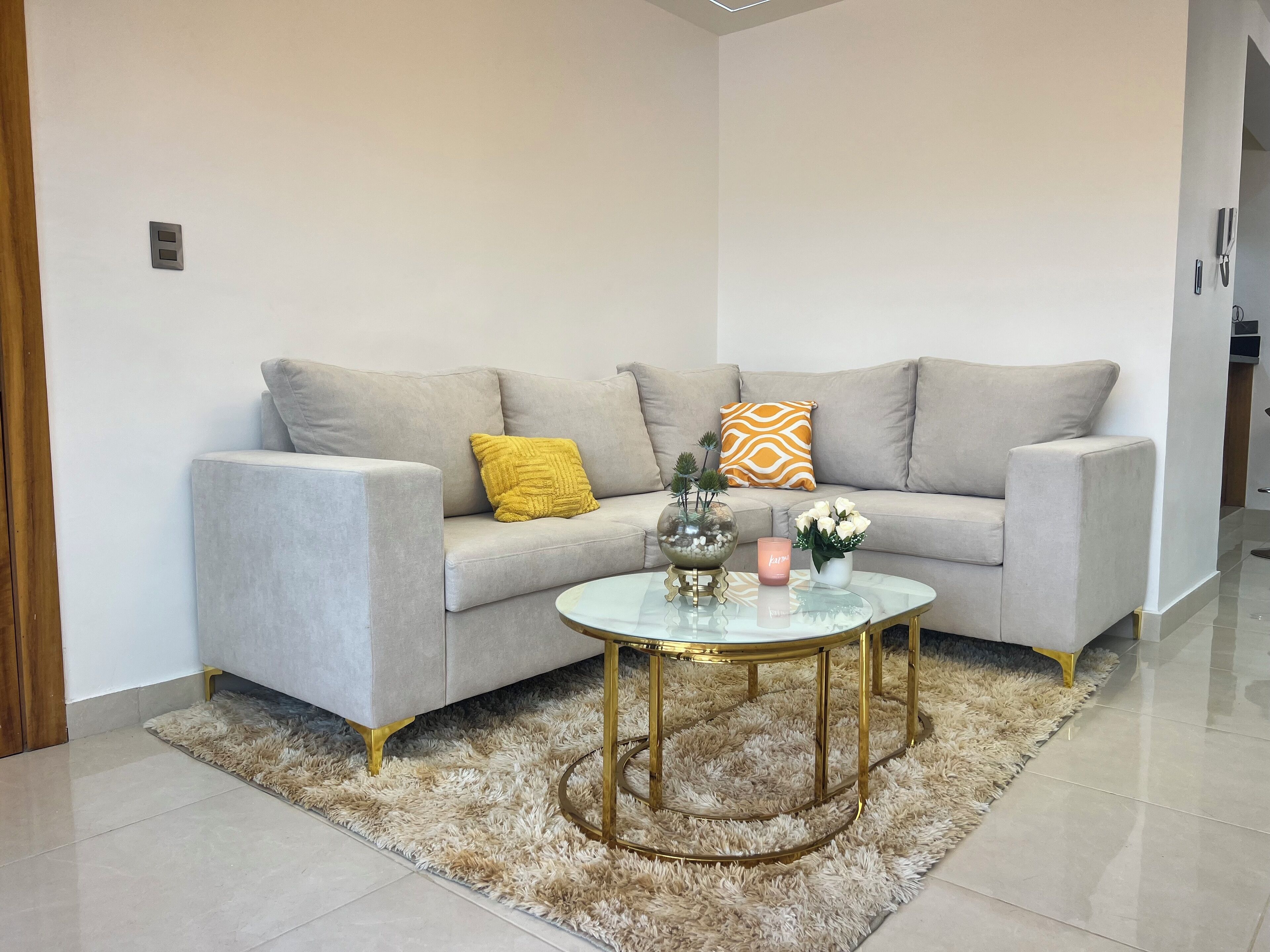 A modern living room with a grey sofa, a glass coffee table, and decorative items including a yellow pillow, a vase with white flowers, and a pink candle.