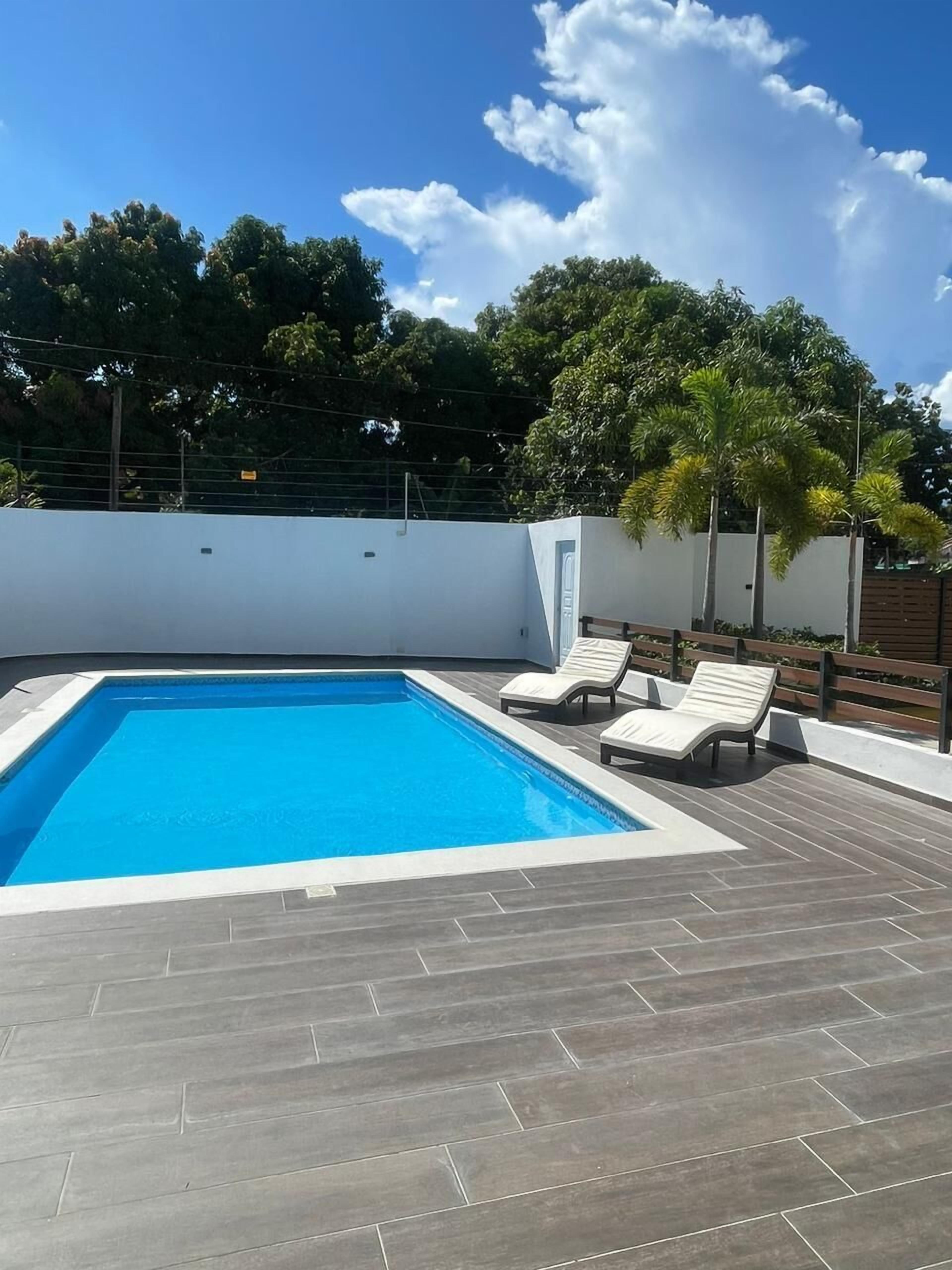 Ein rechteckiger Pool mit Fliesen umgeben, zwei Liegestühle und eine weiße Wand im Hintergrund.